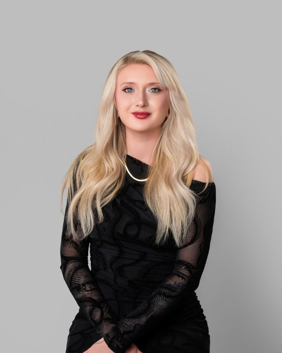 A young woman with long blonde hair, wearing a black off-the-shoulder dress with lace details, and a gold necklace, posing against a plain gray background.