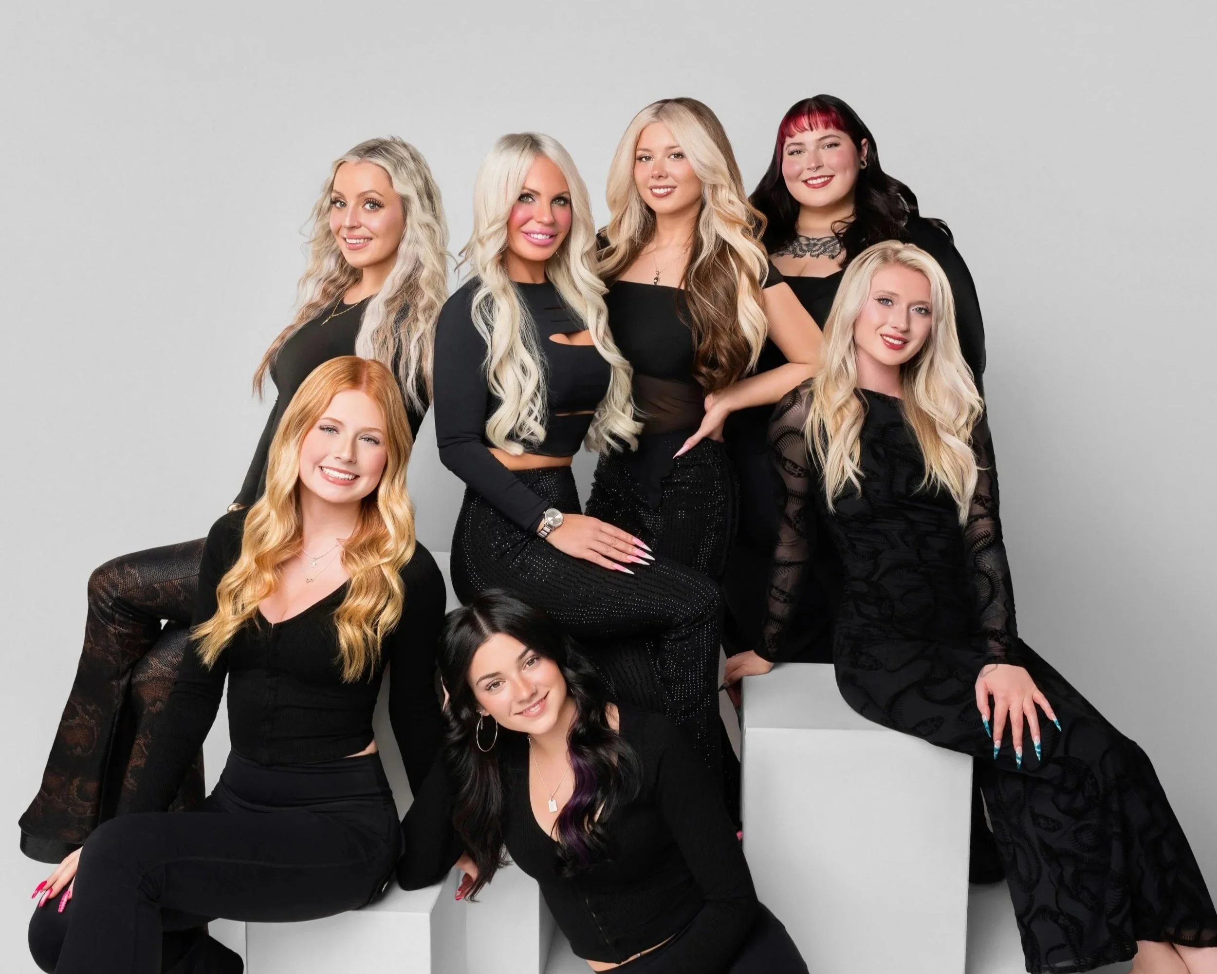 Group of six women with blonde, brunette, red, and black hair, wearing black outfits, posing together against a grey background.