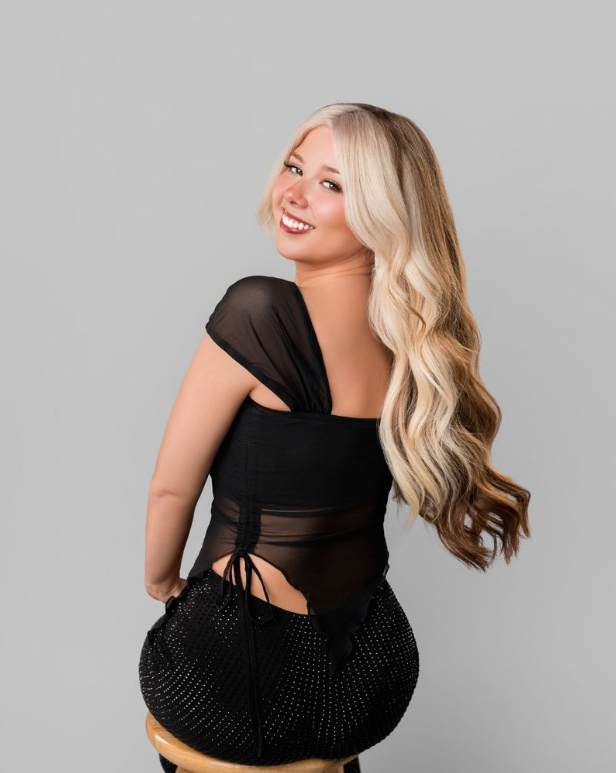 A young woman with long wavy blonde hair, smiling and looking over her shoulder, wearing a black sheer top and black pants with small dots, sitting on a stool against a gray background.