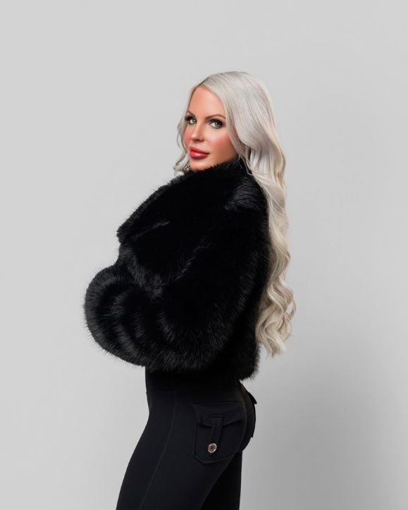 A woman with long, wavy platinum blonde hair wearing a black fur coat and black pants, standing in front of a gray background.