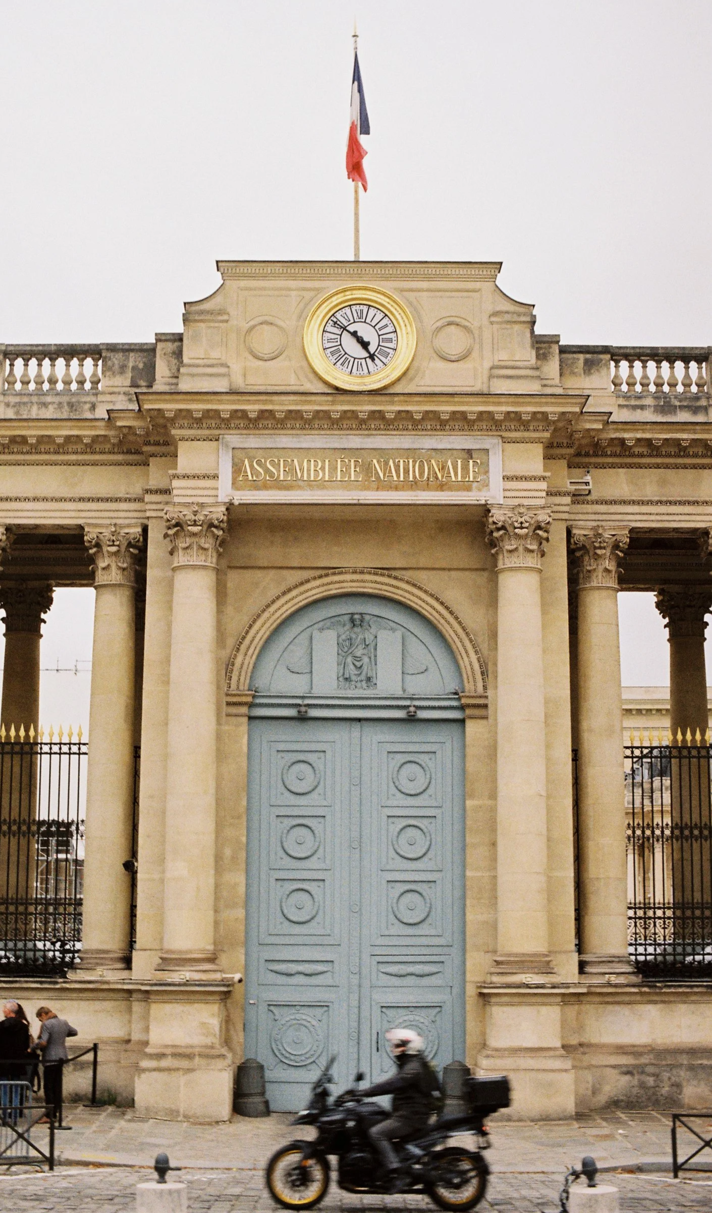 Front view of the entrance to the Assemblée Nationale building with tall columns, a large blue door, a clock showing 5:15, and a French flag on top.