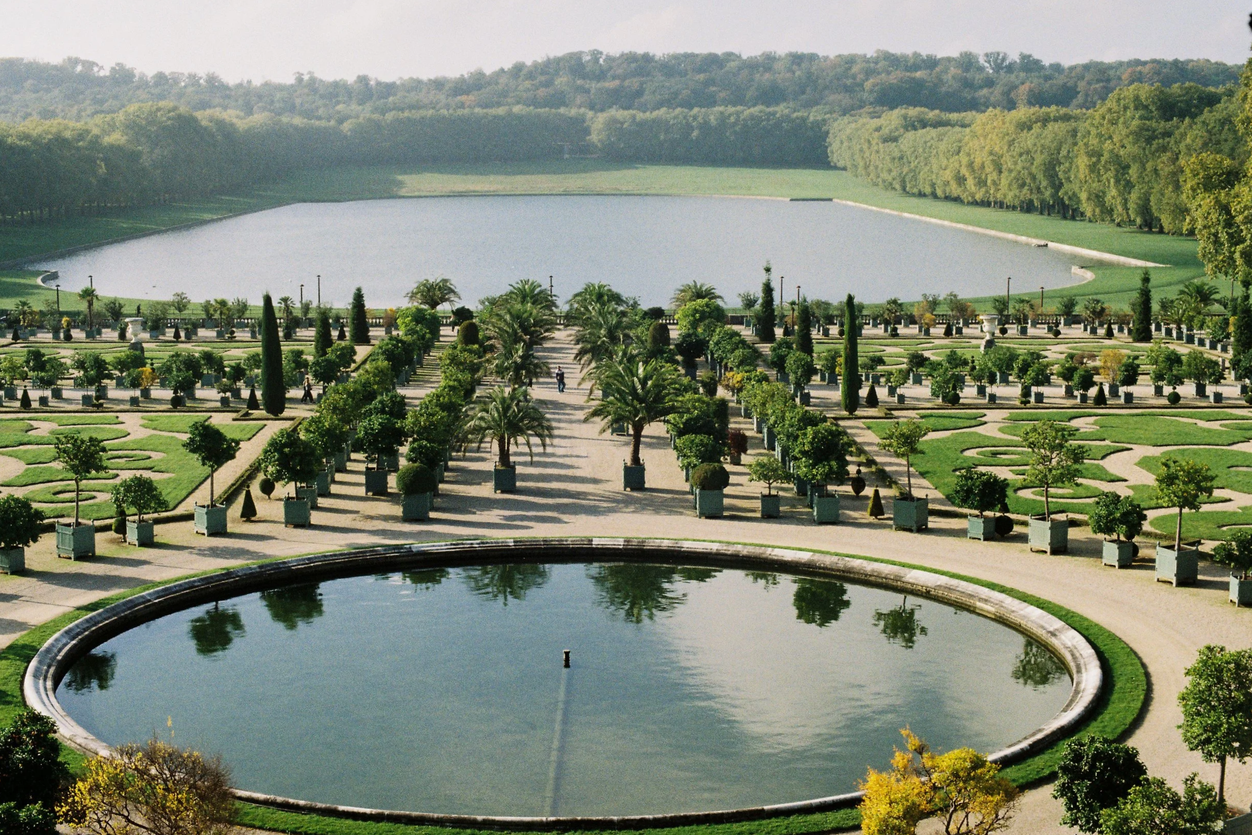 A large, oval-shaped reflective pond surrounded by a formal garden with neatly trimmed trees, shrubs, and paved walkways, with a dense forested hillside in the background.