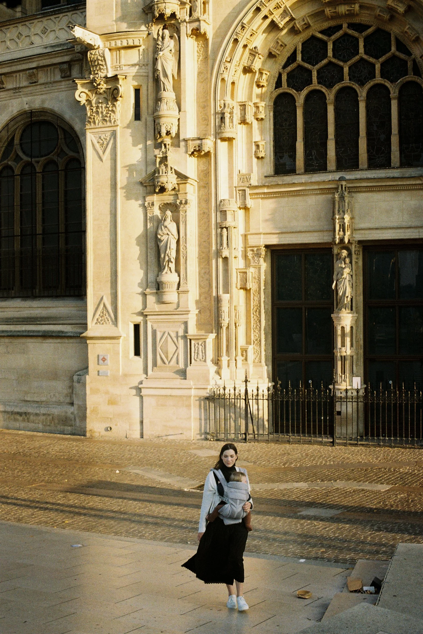 A woman carrying a baby in a carrier walking in front of a historic building with intricate stone carvings and statues, during sunset.