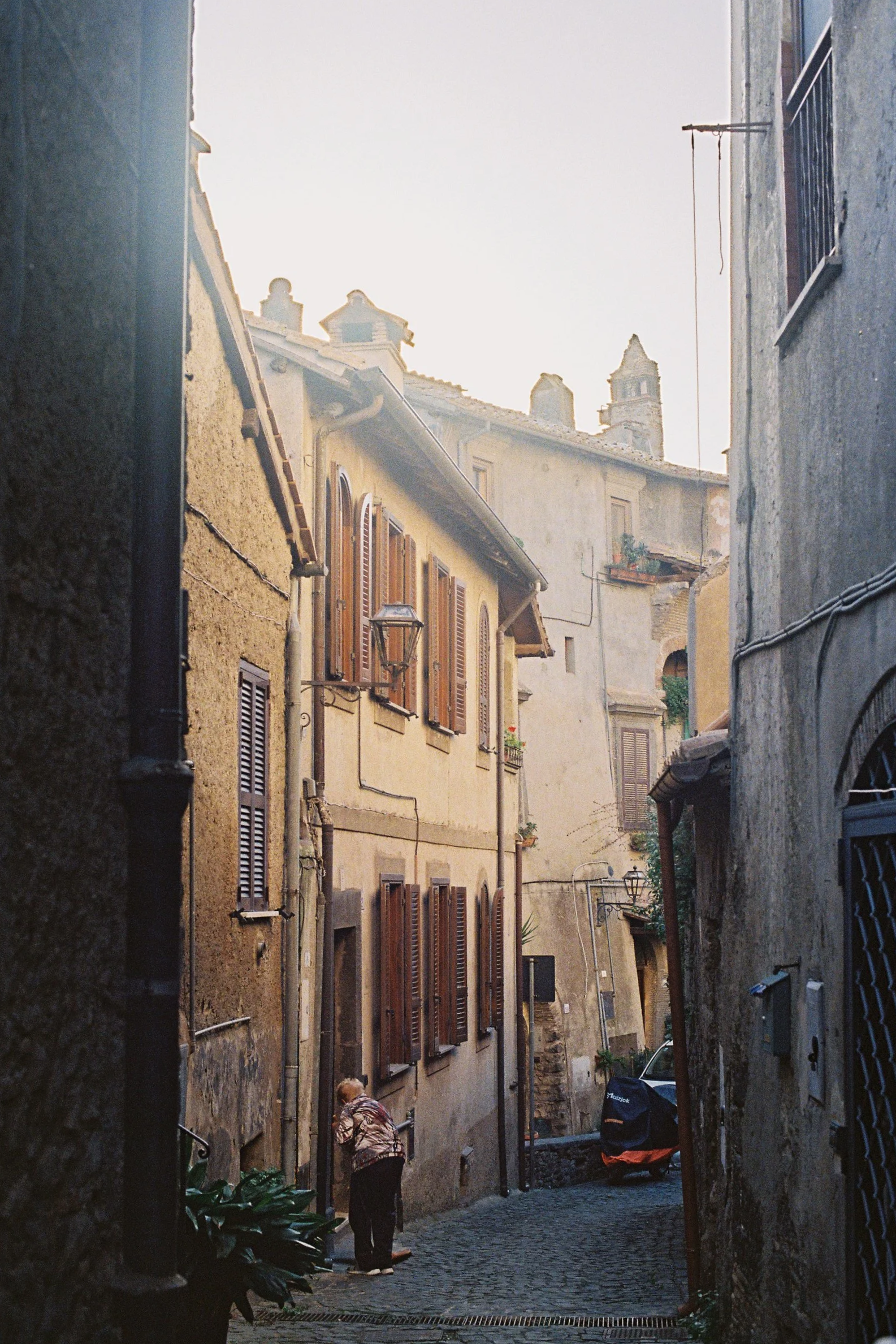 A narrow cobblestone street in an old European town, with closely packed buildings on both sides, shuttered windows, and a person walking while another is bent over near some plants.