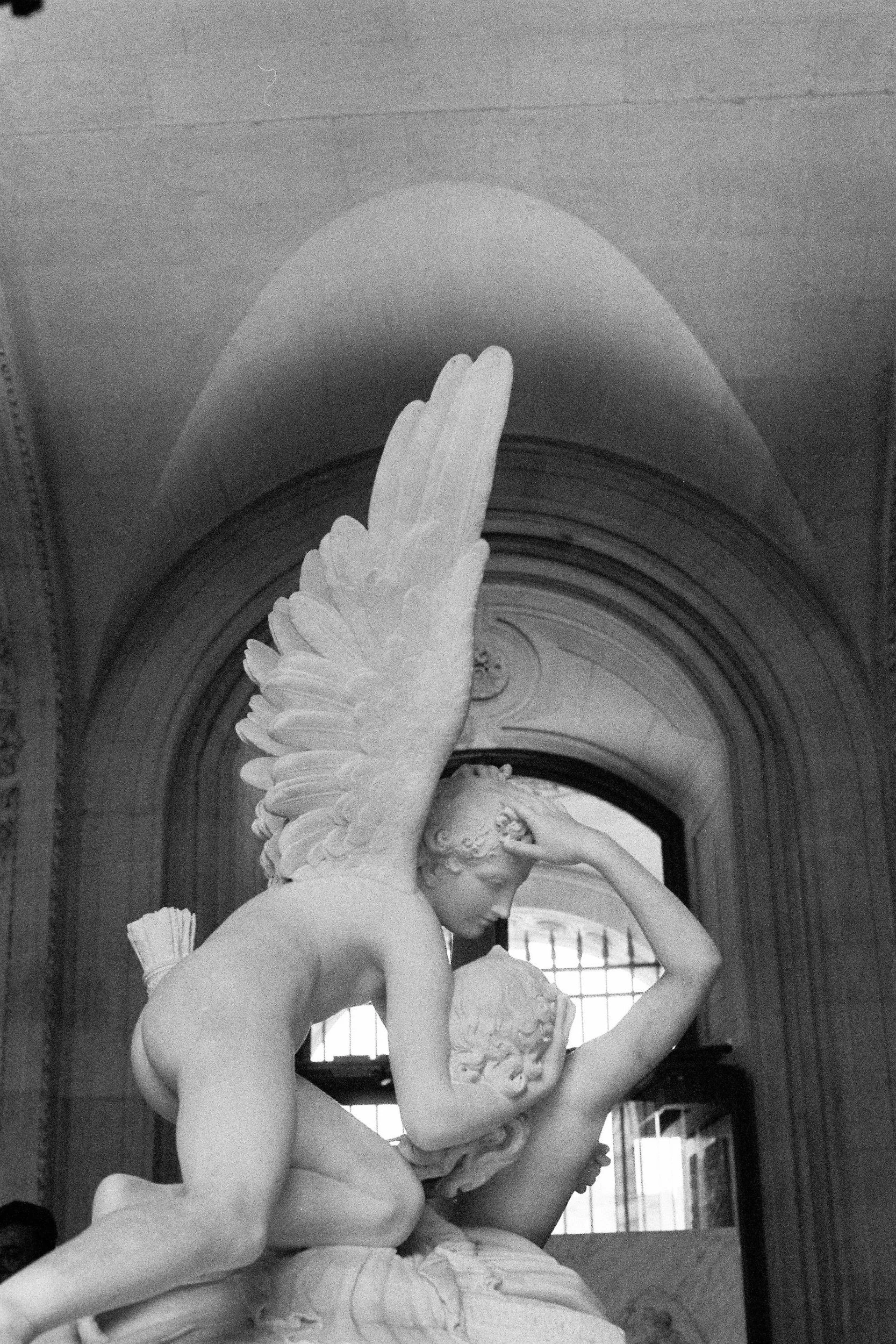 A black and white photo of a marble sculpture of a woman with large wings, standing in front of a window with bars, inside a building.