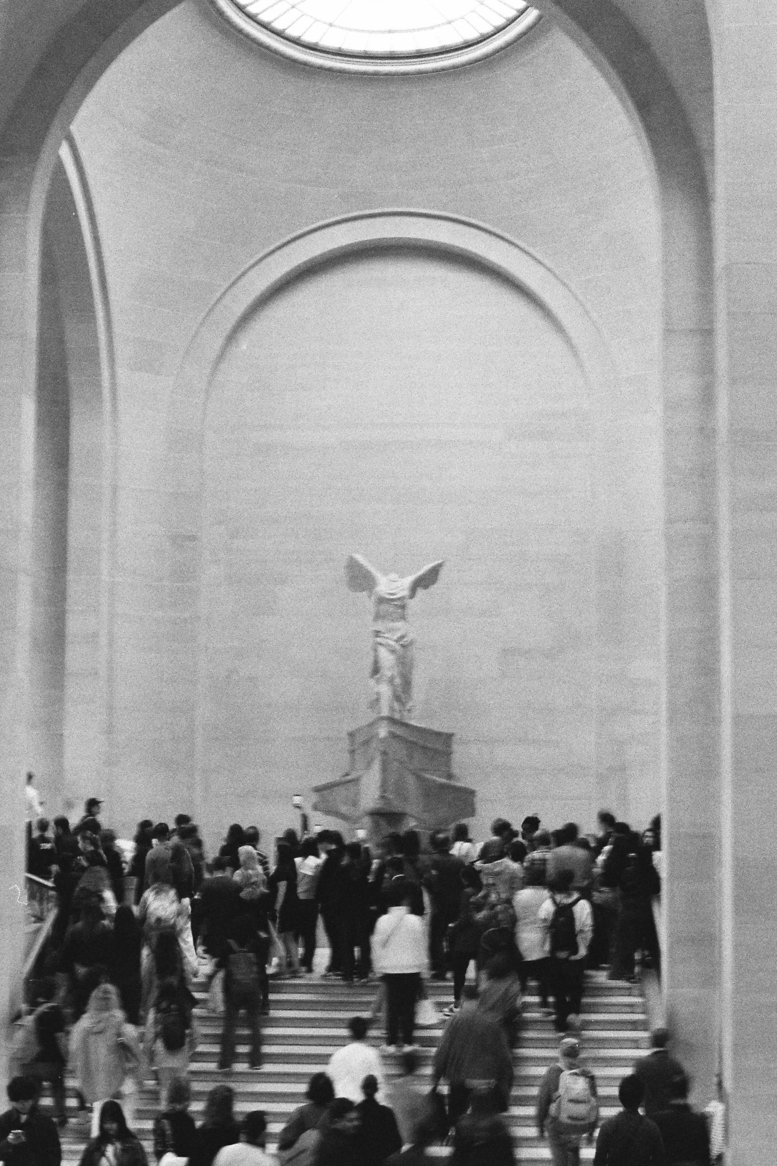 Black and white photo of a large crowd of people ascending stairs and gathering in a grand interior space. A statue of a winged figure stands on a pedestal in the center background, and an arched ceiling is visible above.