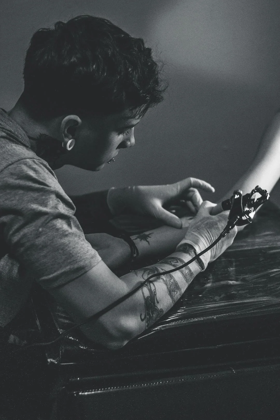 A person with tattoos and gauges in their ears is sitting at a table, looking down at their phone, in a black and white photo.