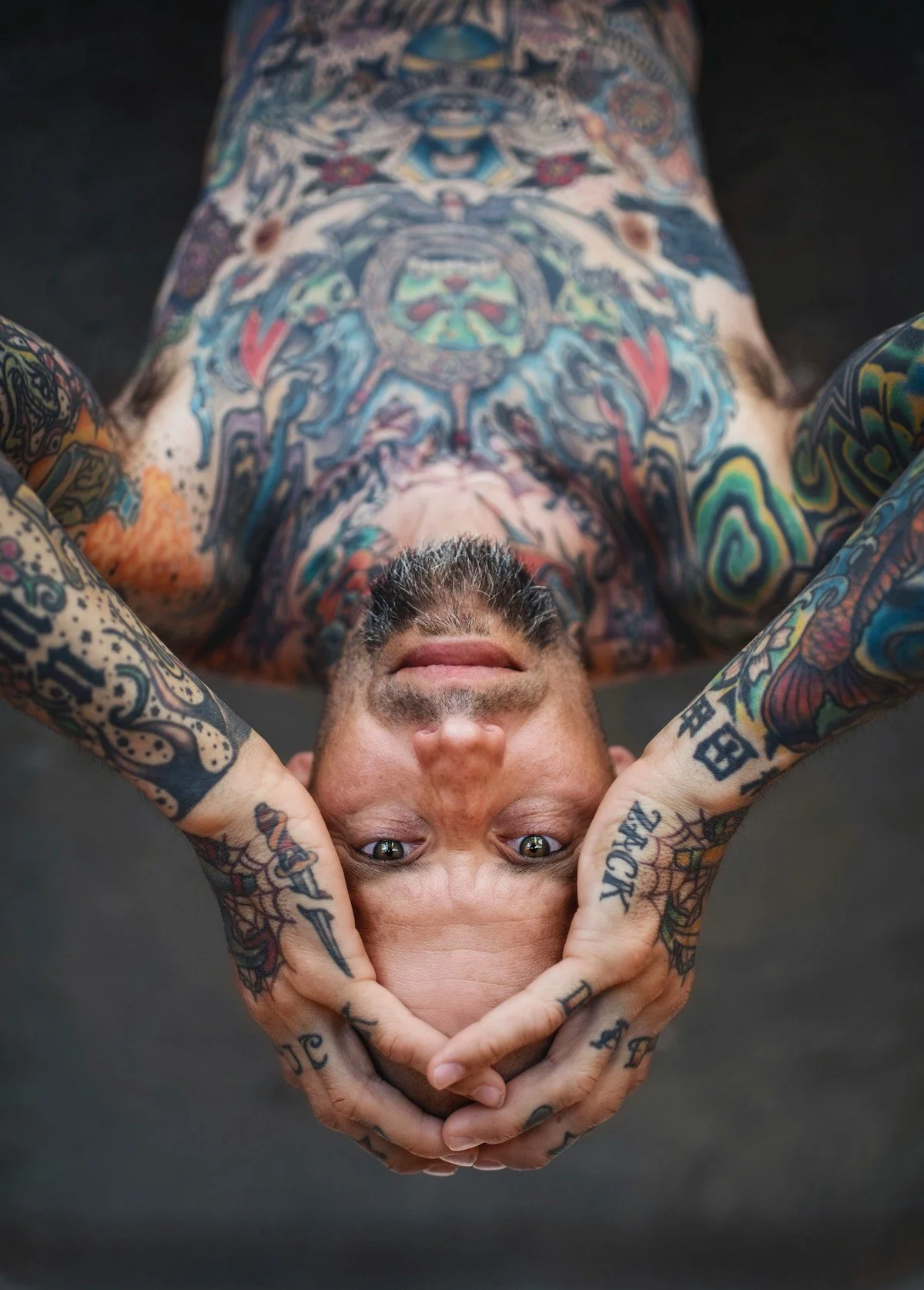 Tattooed man lying on his back with hands framing face, looking up at the camera.