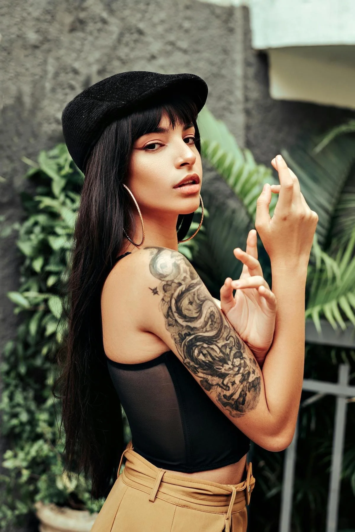 A woman with long dark hair, wearing a black beret and hoop earrings, has a large tattoo on her left arm and is posing outdoors in front of greenery and a textured wall.