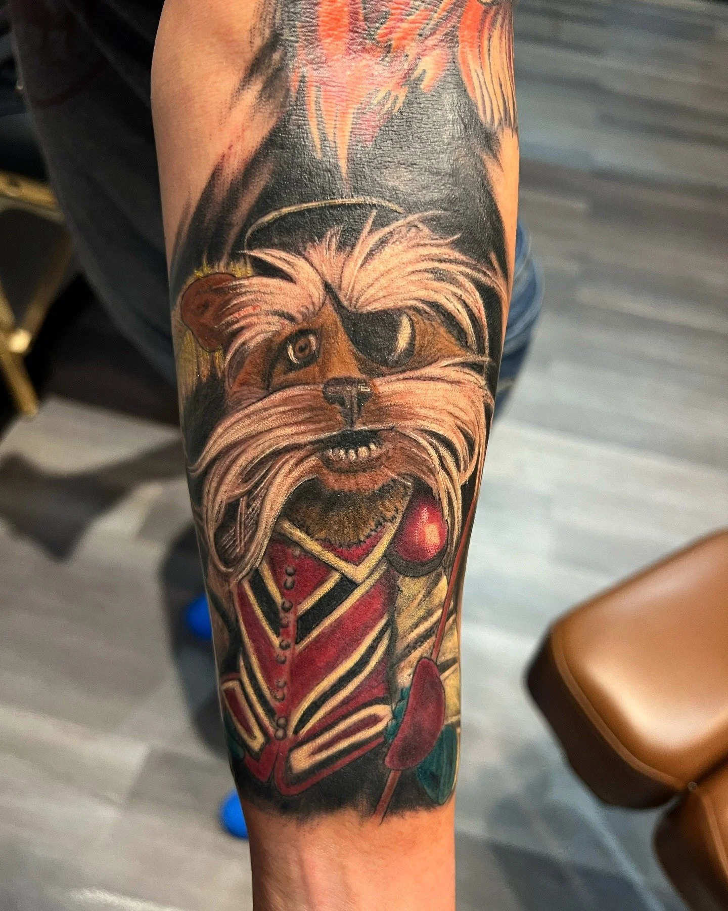 Tattoo of a dog with a pirate patch and an eye patch, dressed in a red and gold regal outfit, holding a sword on a person's forearm.