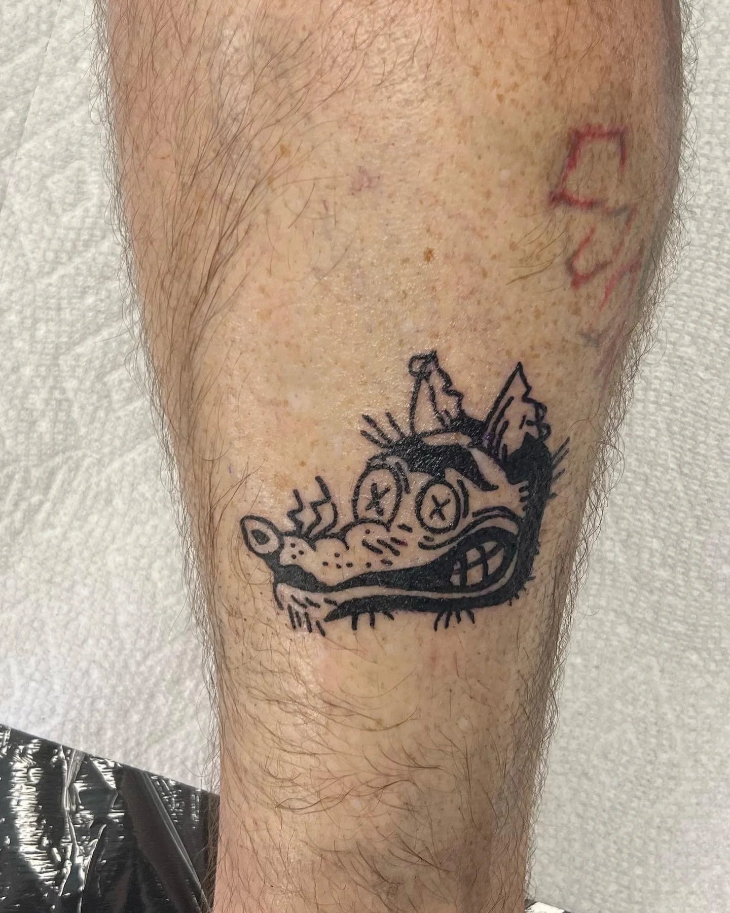 A tattoo of a cartoonish creature with exaggerated features, including large crossed-out eyes, a wide open mouth with teeth, a spotted nose, and two parts that resemble ears or horns, on a person's leg.