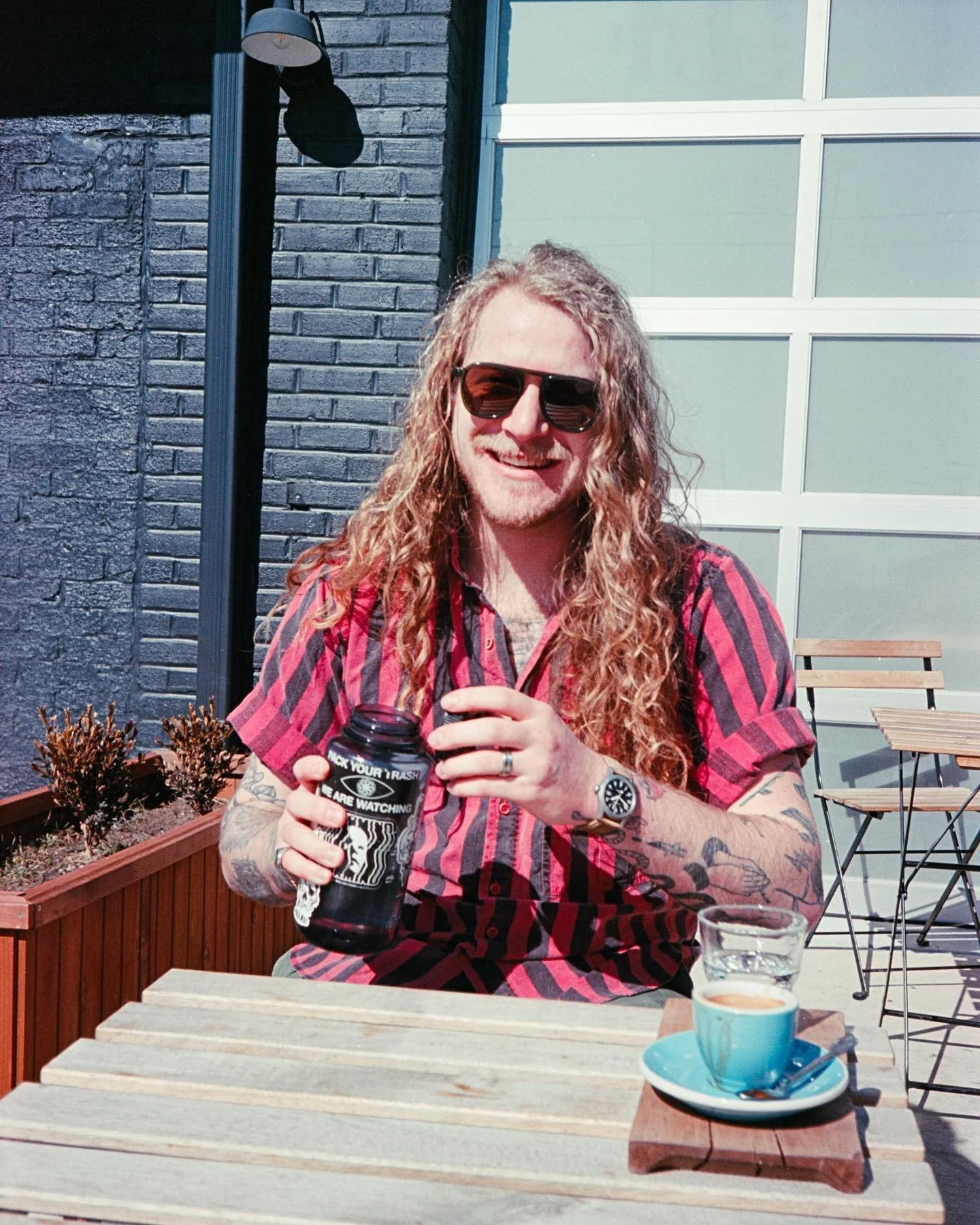 A man with long curly hair, tattoos on his arms, wearing sunglasses, a red and black checkered shirt, smiling while holding a dark bottle, sitting at an outdoor patio table with a blue cup of what appears to be coffee or tea in front of him.