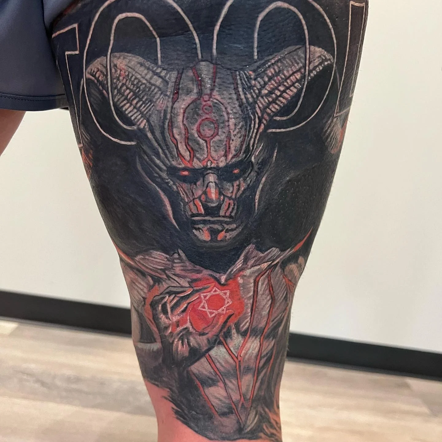 Tattoo of a demon with horns, dark eyes, and a skeletal face, holding a glowing red heart with a pentagram symbol, covering the thigh.