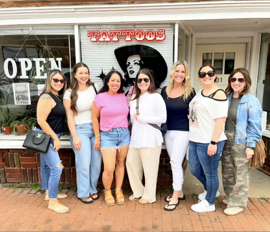 Group of eight women standing outside a tattoo shop, smiling, with the shop's sign reading 'TATTOOS' and an 'OPEN' sign in the window.