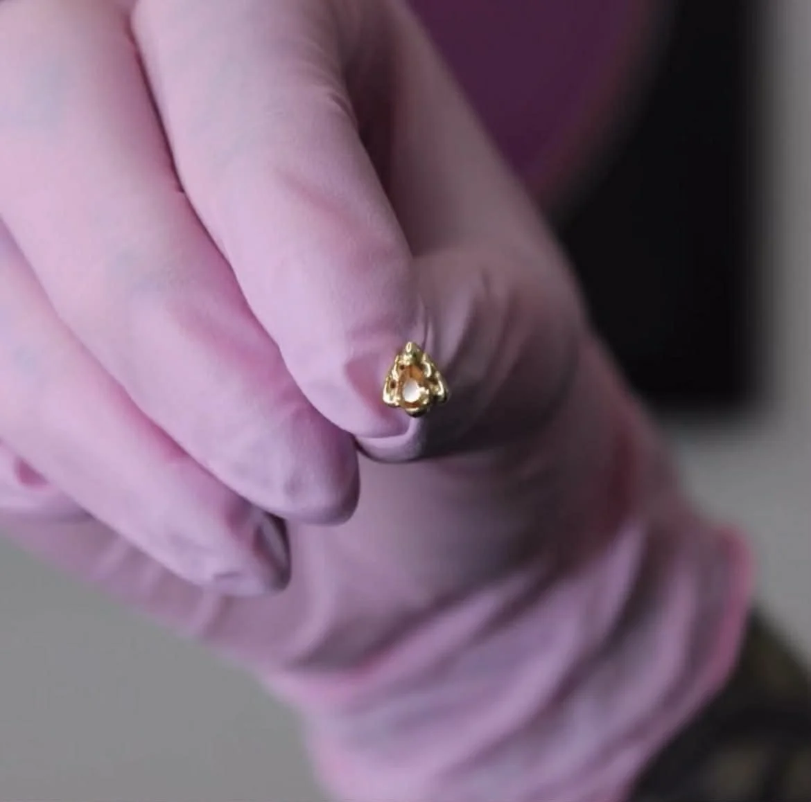 A person wearing a pink glove holding a gold ring with a heart-shaped design.