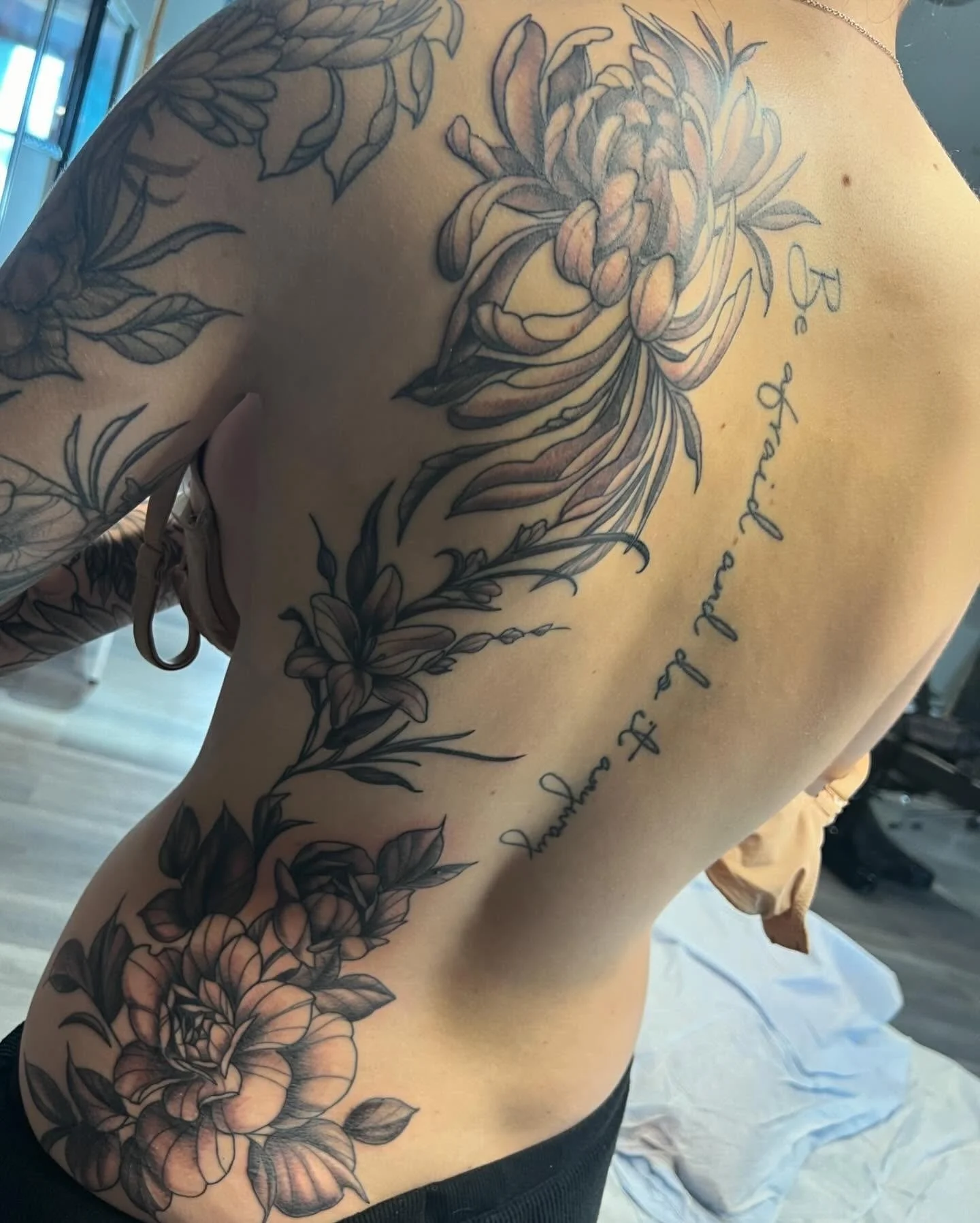 A person's back with a large floral tattoo and cursive text. The tattoo includes chrysanthemums and lilies, and the text reads 'Be Knash and Jaccurs of F'.