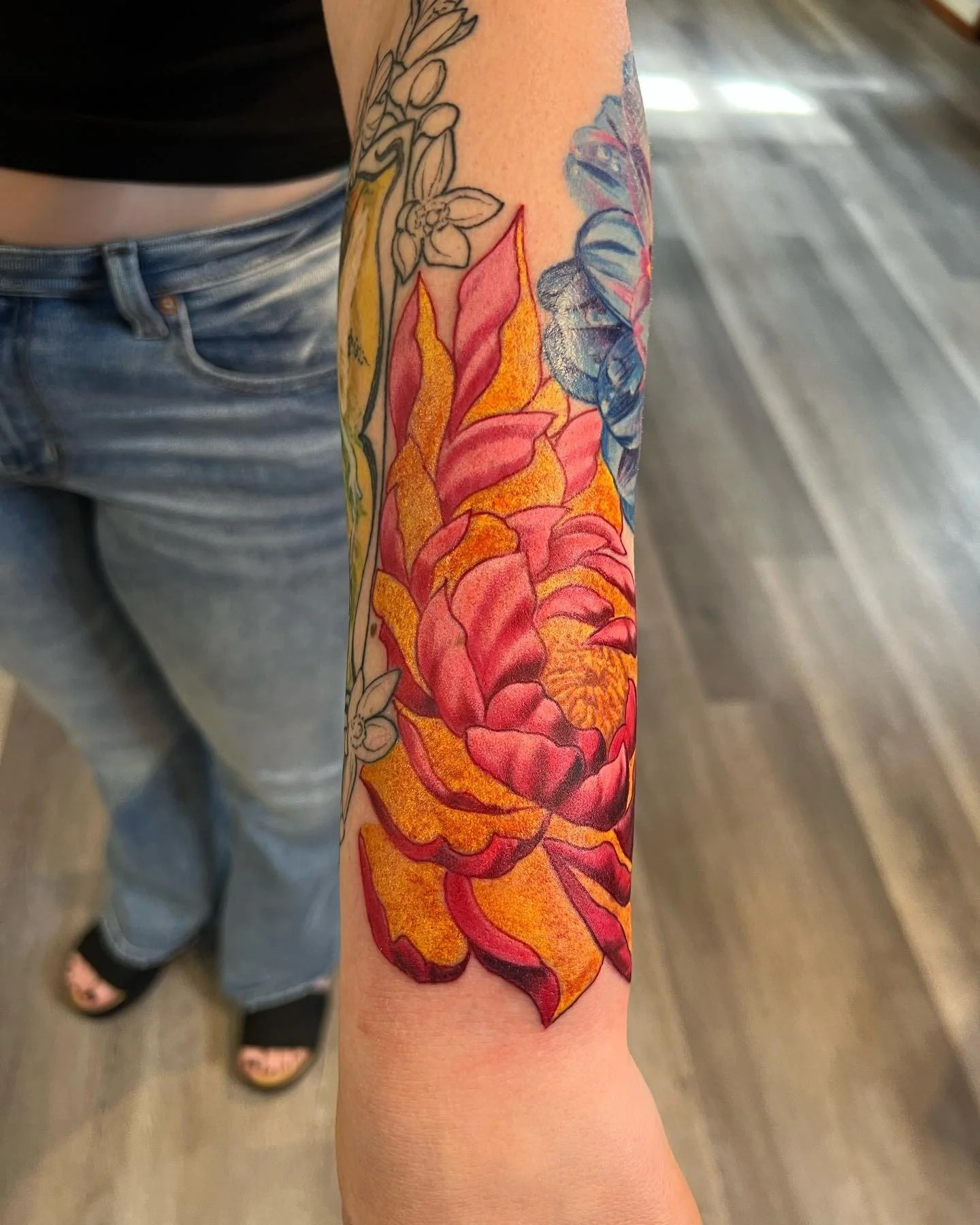 Colorful floral tattoo on a person's forearm featuring a large pink and orange flower with green leaves.