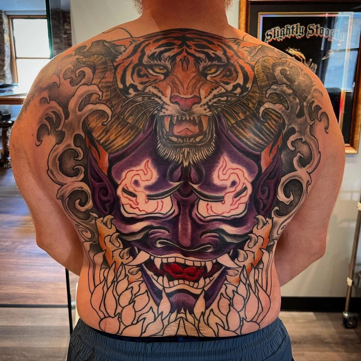 Colorful tattoo of a fierce tiger on the person's upper back and shoulders, with a stylized face below. The tiger has sharp teeth, vivid orange and black stripes, and a pink nose.
