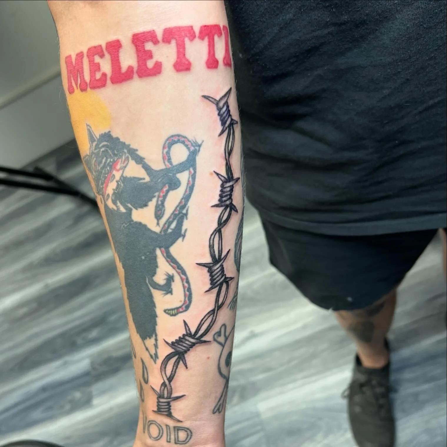 Tattoo on person's forearm featuring the word 'Melett' in red, a black lion holding a snake, and a barbed wire design with the words 'No Kid' partially visible.