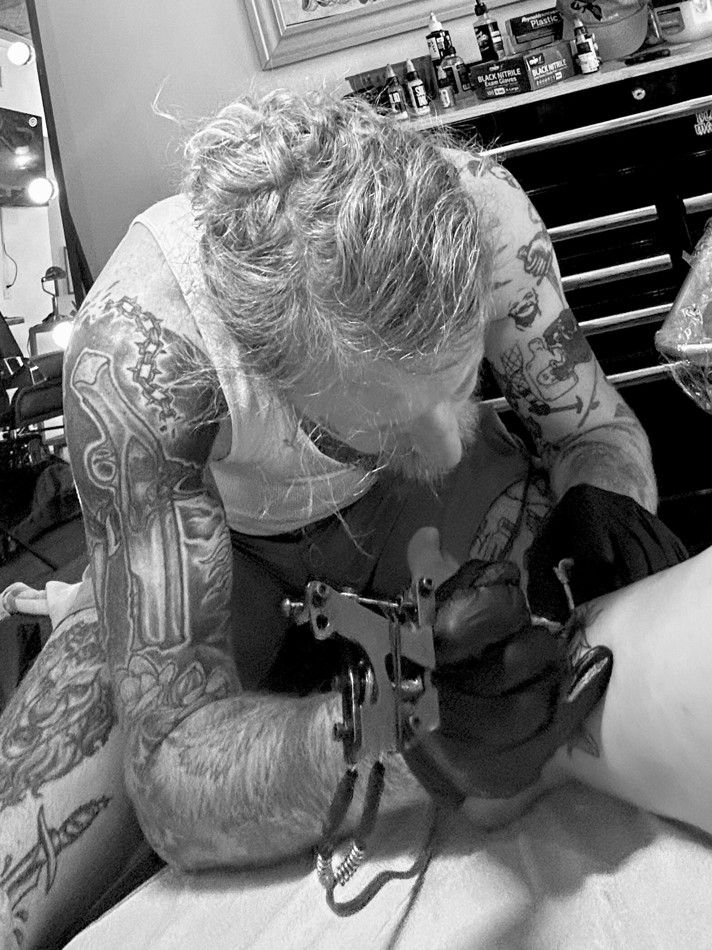 A tattooed man with long hair is tattooing a woman's leg at a tattoo parlor. The man is wearing black gloves and is focused on his work. Various tattoo supplies and bottles are visible on the shelves in the background.