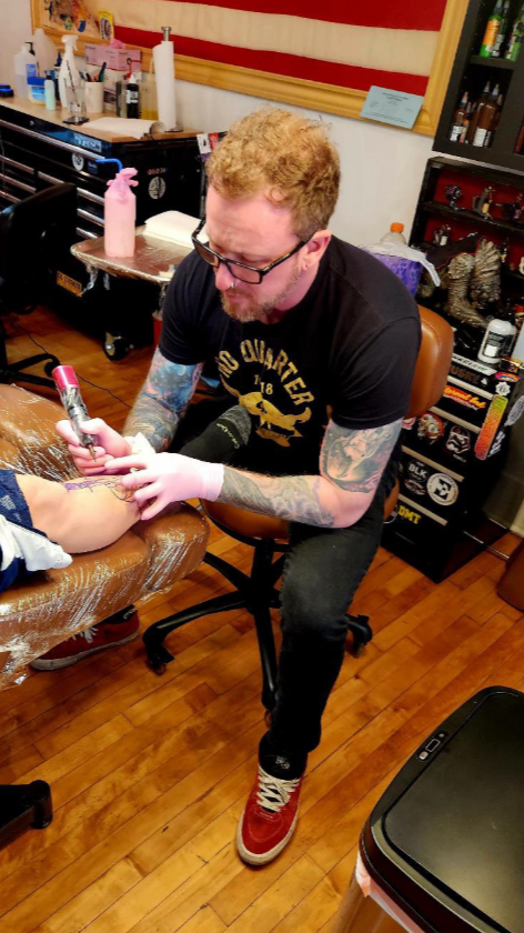 A man with red hair and glasses getting a tattoo on his arm in a tattoo parlor.