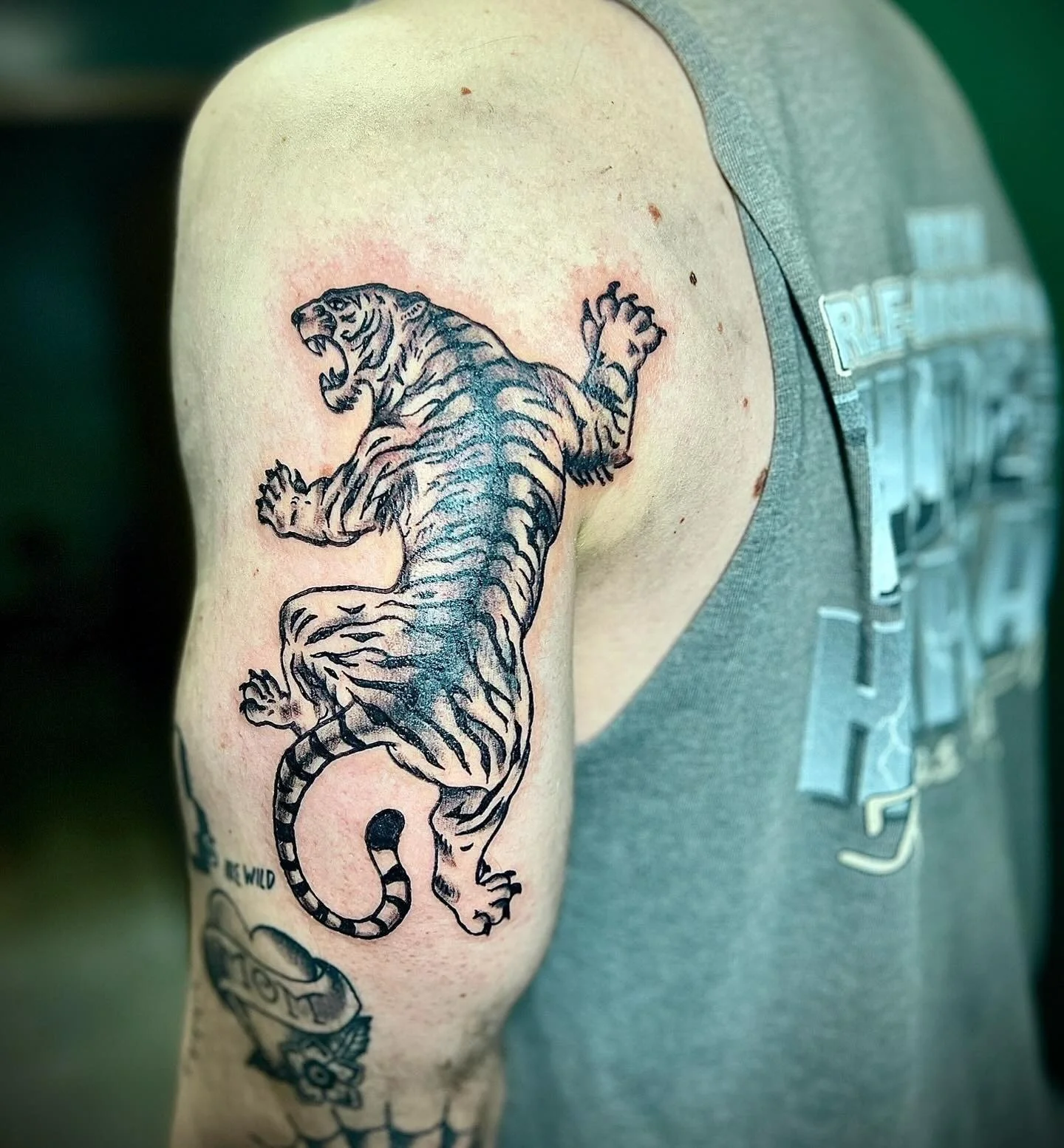Tattoo of a roaring tiger on upper arm of person wearing a gray tank top.