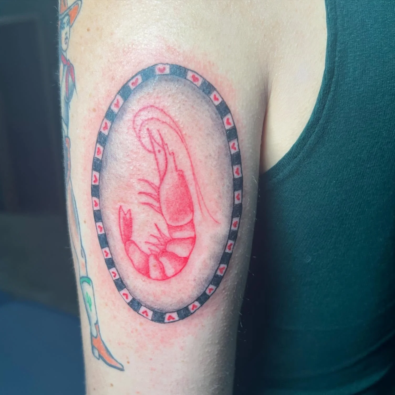 Tattoo of a red outline of a shrimp in an oval frame with a playing card pattern, located on a person's upper arm.