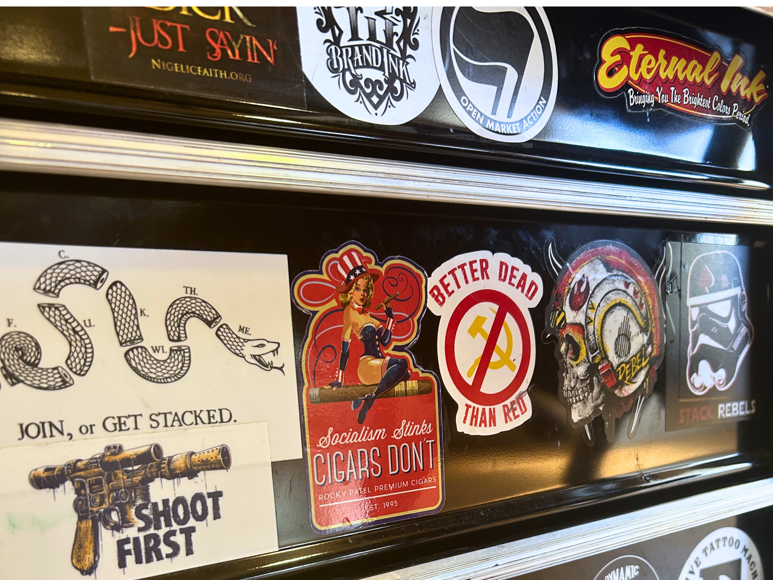 Collection of various stickers on a metallic surface, including a peace sign with a woman holding a cigarette, a skull with headphones, a Star Wars stormtrooper helmet, and a vintage-style poster of a snake with text, alongside some other patches and stickers.