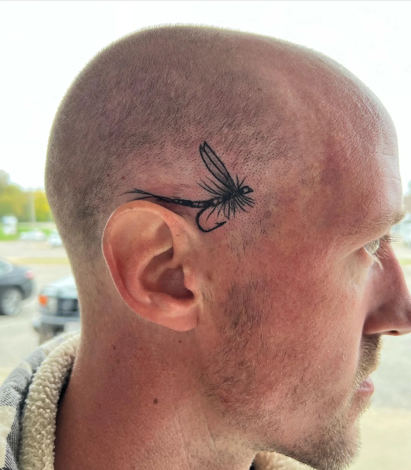 A man with a and buzz haircut reveals a tattoo of a dragonfly on the side of his head above his ear