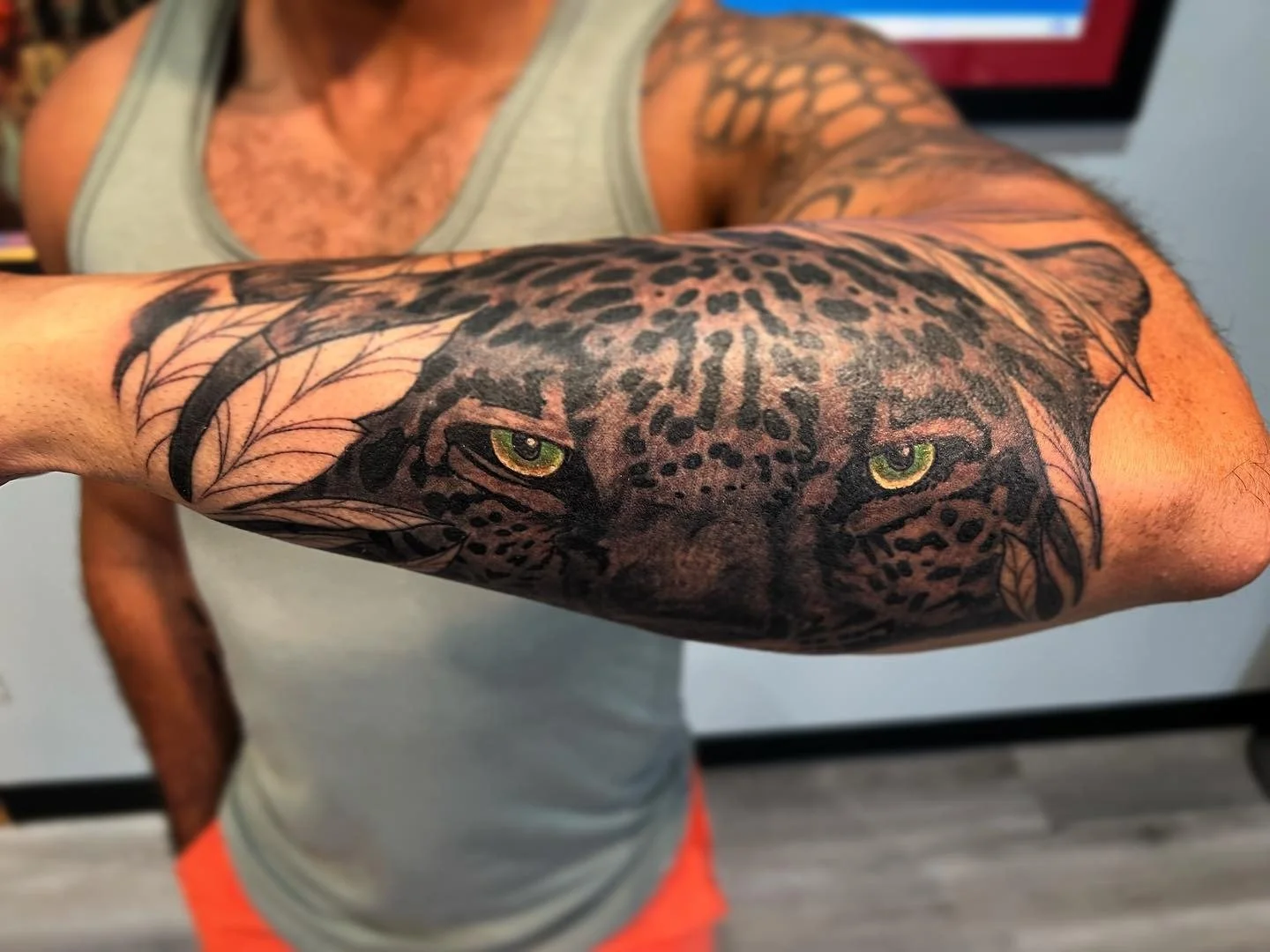 Tattoo of a realistic leopard face with piercing yellow eyes on a person's arm, surrounded by leaf designs.