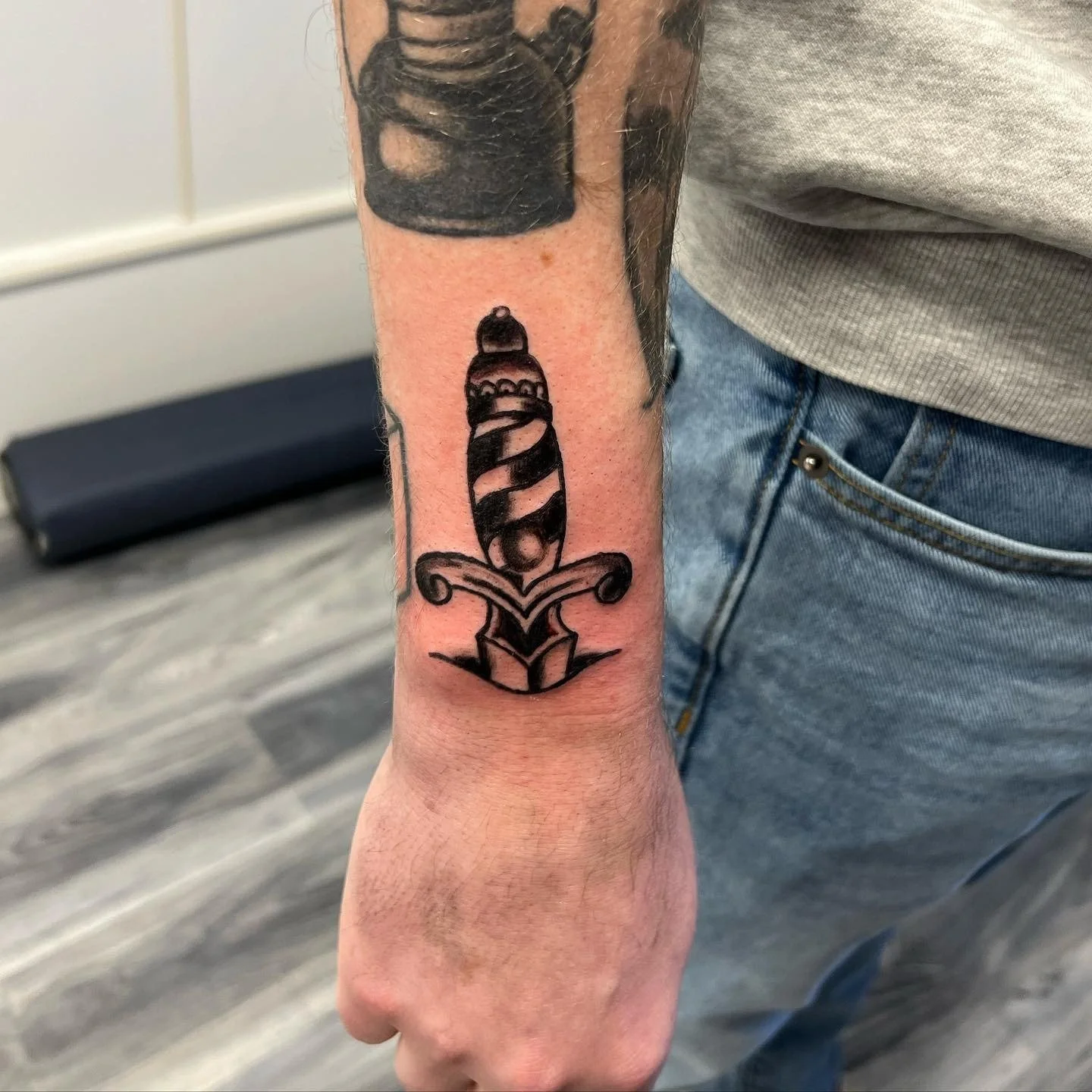 Tattoo of a dagger with a striped handle, pointed downward, on a person's inner forearm.