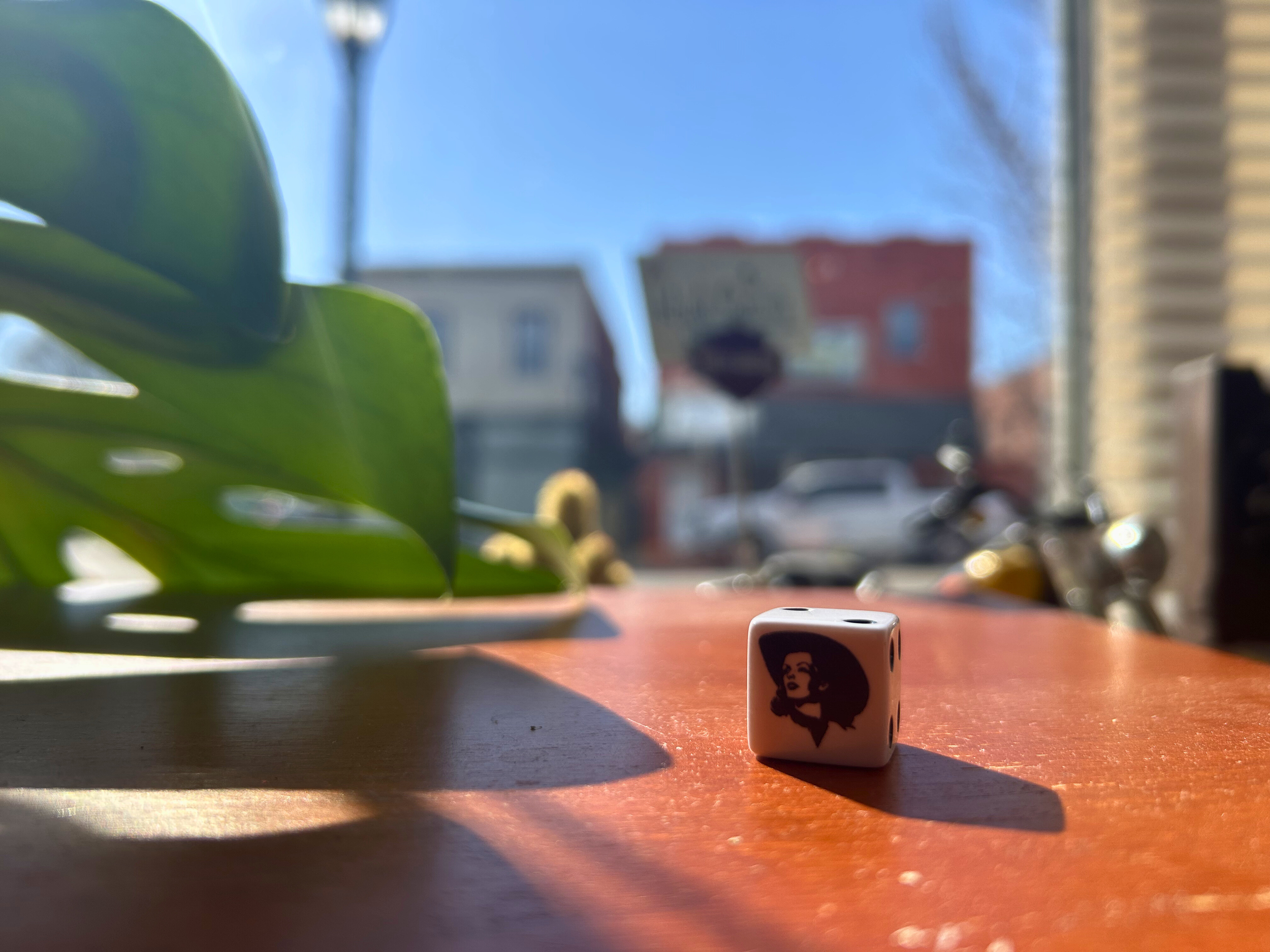 A small die featuring a woman's face with a large hairdo and a confident expression. The die is placed on a wooden surface near a green plant and sunlight, with a blurred background of buildings, a street sign, and parked bikes outside.