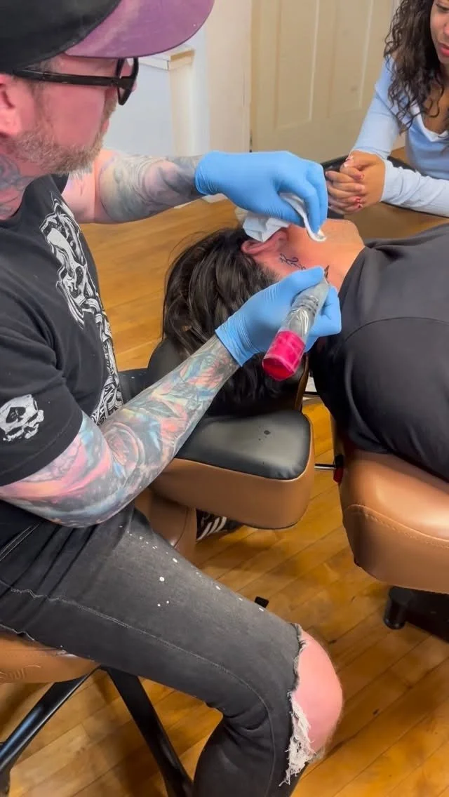 Tattoo artist tattooing a person's neck in a tattoo studio, with another person observing.