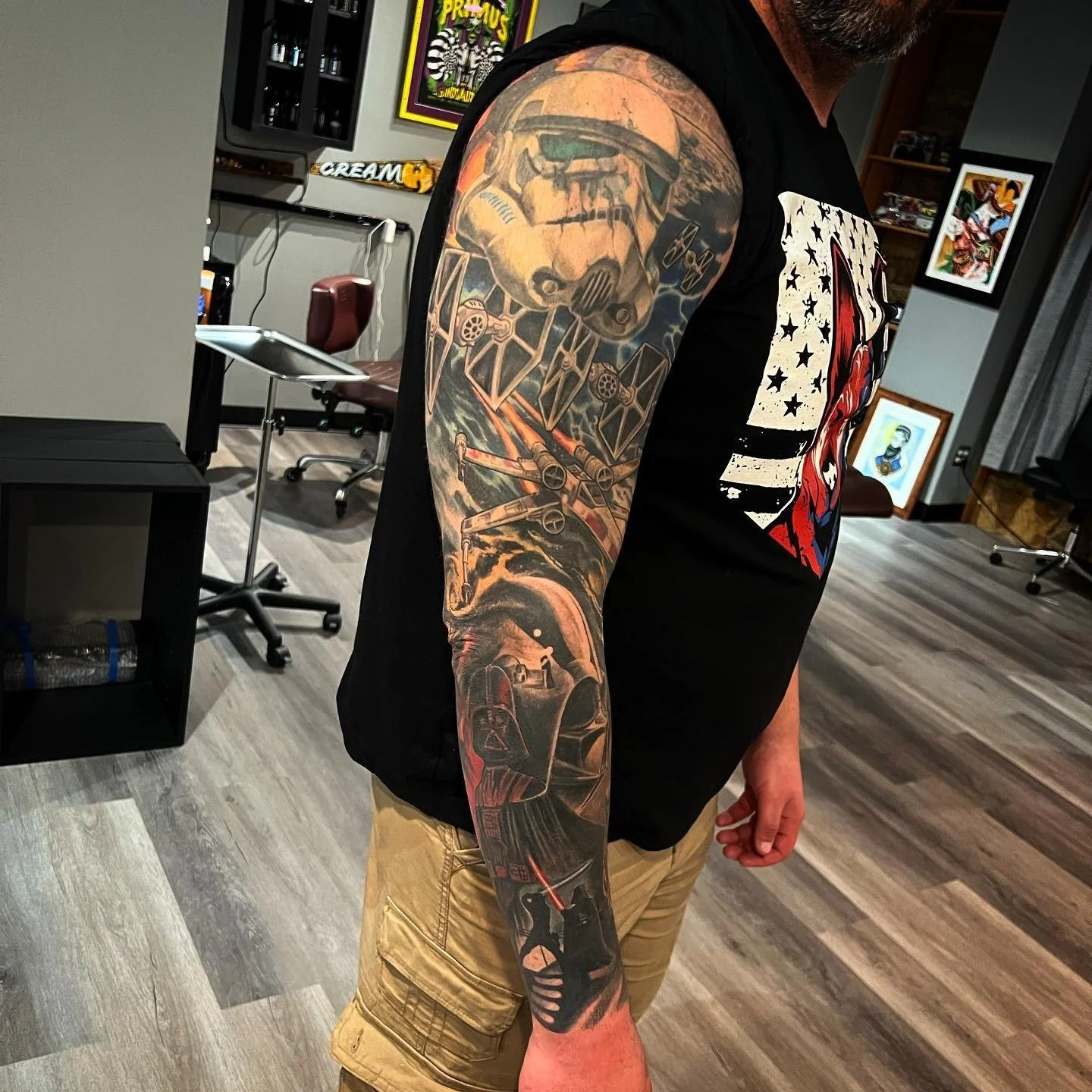 A man with a large Star Wars-themed tattoo sleeve on his left arm, featuring a Stormtrooper helmet, TIE fighters, and Darth Vader, is standing indoors in front of artwork and furniture.