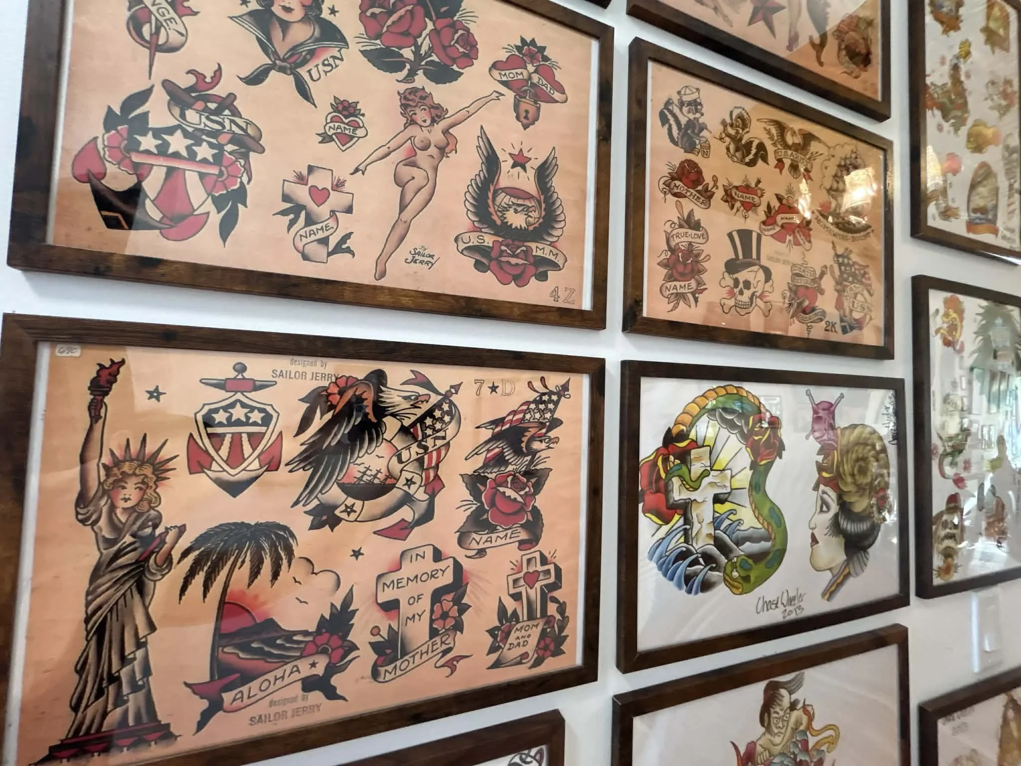 Collection of framed traditional tattoo flash designs on a wall, featuring symbols including eagles, roses, skulls, a snake, and various patriotic themes.