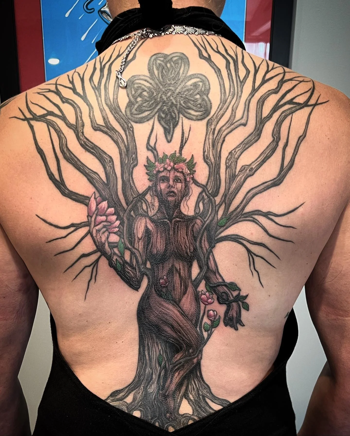 A large tattoo on a person's back depicting a tree with human features, blending into a figure of a woman with flowers in her hair, surrounded by pink roses and green leaves, with a large four-leaf clover at the top.