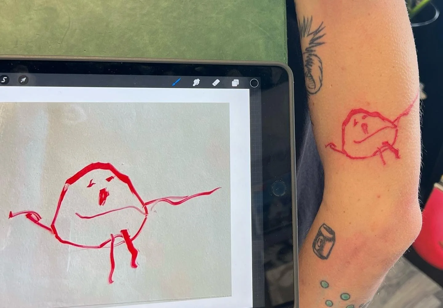 A tattoo of a simple, childlike red line drawing of a face on a person's forearm next to a digital tablet display showing the same drawing.