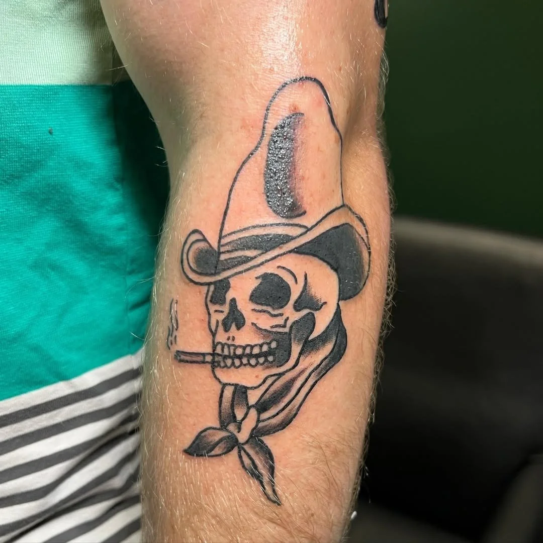 Tattoo of a skull wearing a cowboy hat and smoking a cigarette, with a bandana around its neck.