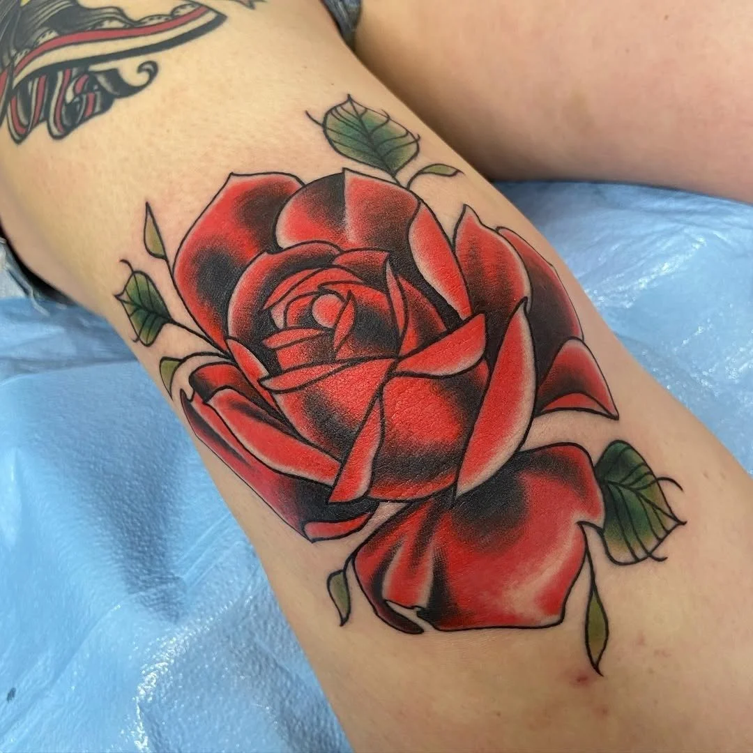 Tattoo of a large red and black rose with green leaves on a person's arm.
