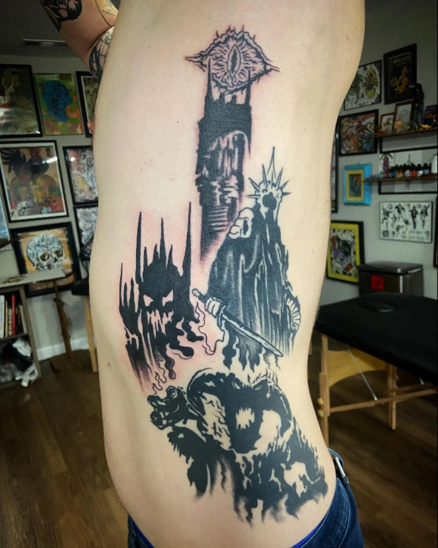 Black tattoo on person's side featuring various dark, fantasy-inspired characters and scenes, including a dragon head, a monstrous creature, a figure wielding a sword, and a tower with a grotesque face at the top.