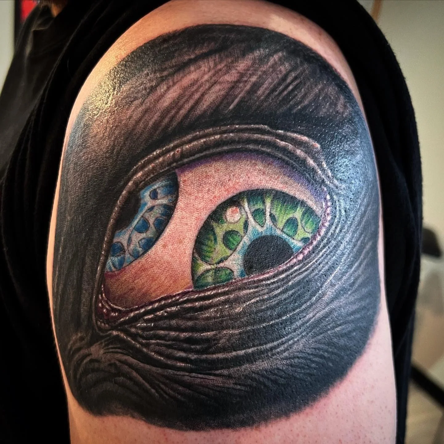 Tattoo of a realistic eye with a vibrant green and blue iris, partially covered by black shading that resembles eyelids or shadows.