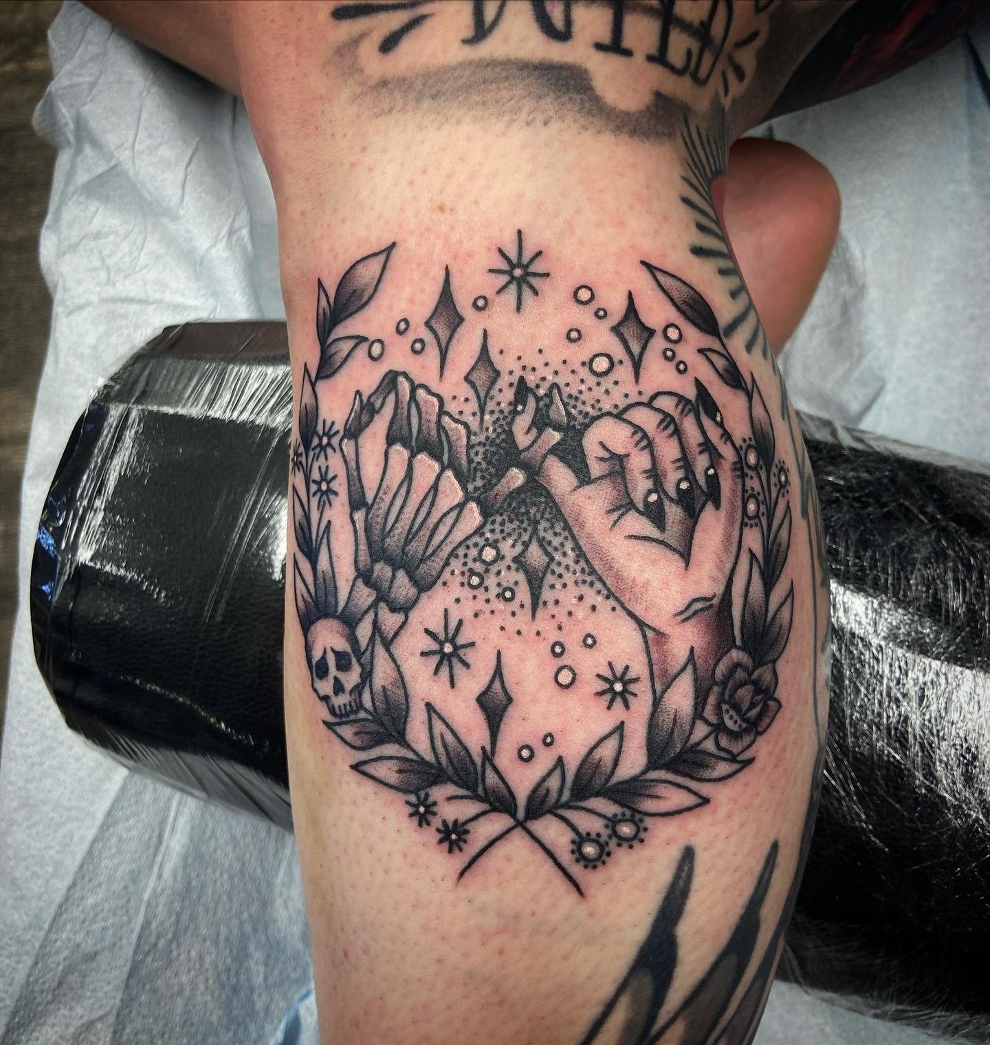 Tattoo on arm featuring a skeleton hand and a human hand making a fist, surrounded by leaves, stars, diamonds, a skull, and abstract patterns.