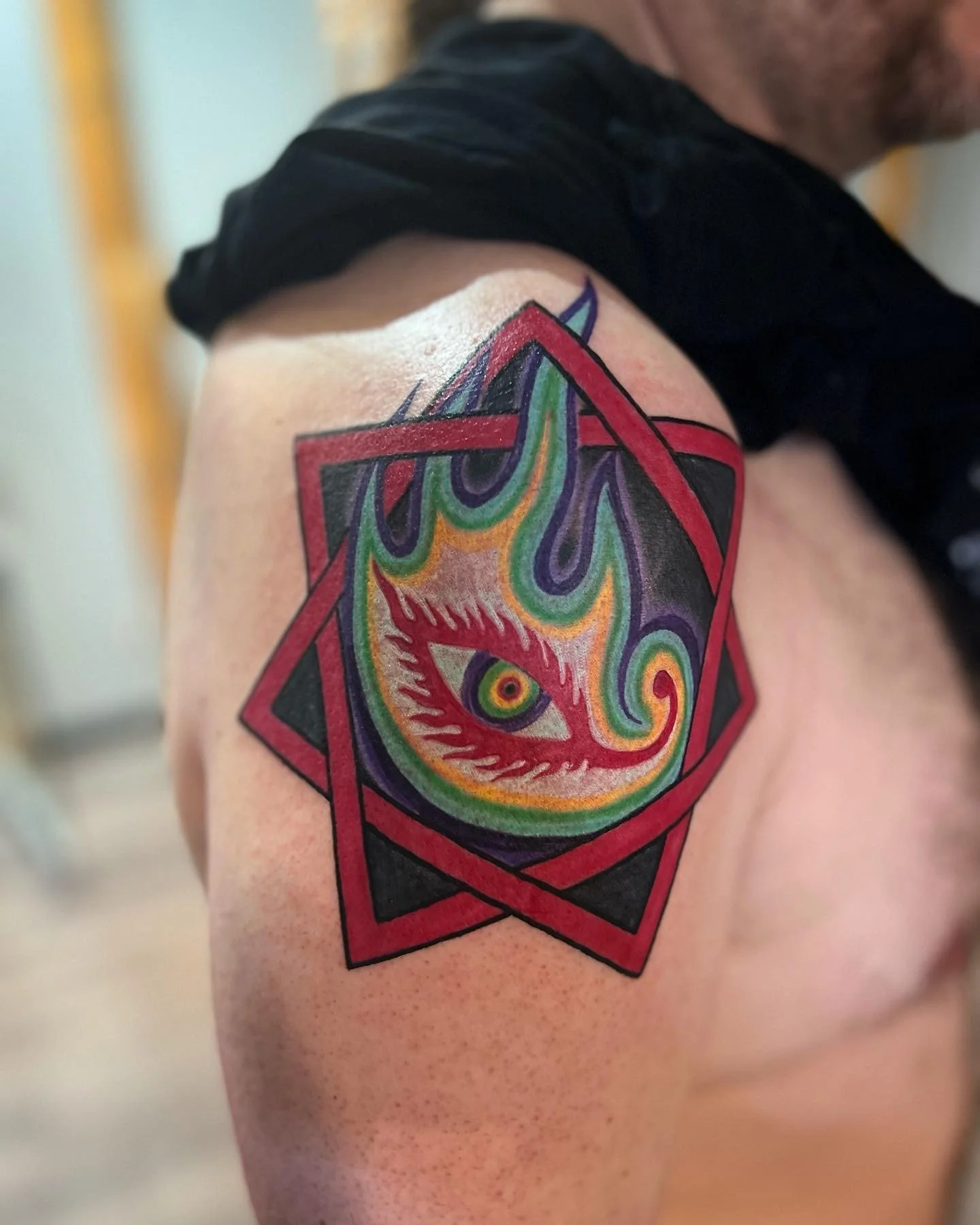 Colorful tattoo of a stylized eye with flames inside a geometric frame on a person's upper arm.