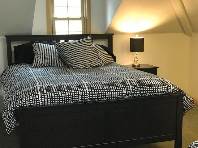 A neatly made bed with black and white checkered bedding, two checkered pillows, a wooden headboard, a bedside table with a black lamp, and a window with a white frame in the background.