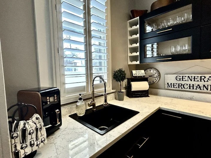 Kitchen counter with a black sink, a soap dispenser, a faucet, a small potted plant, a coffee maker, a paper towel holder, a clock, and wall decor that says "General Merchandise." Includes a window with white shutters and glass-front cabinets.