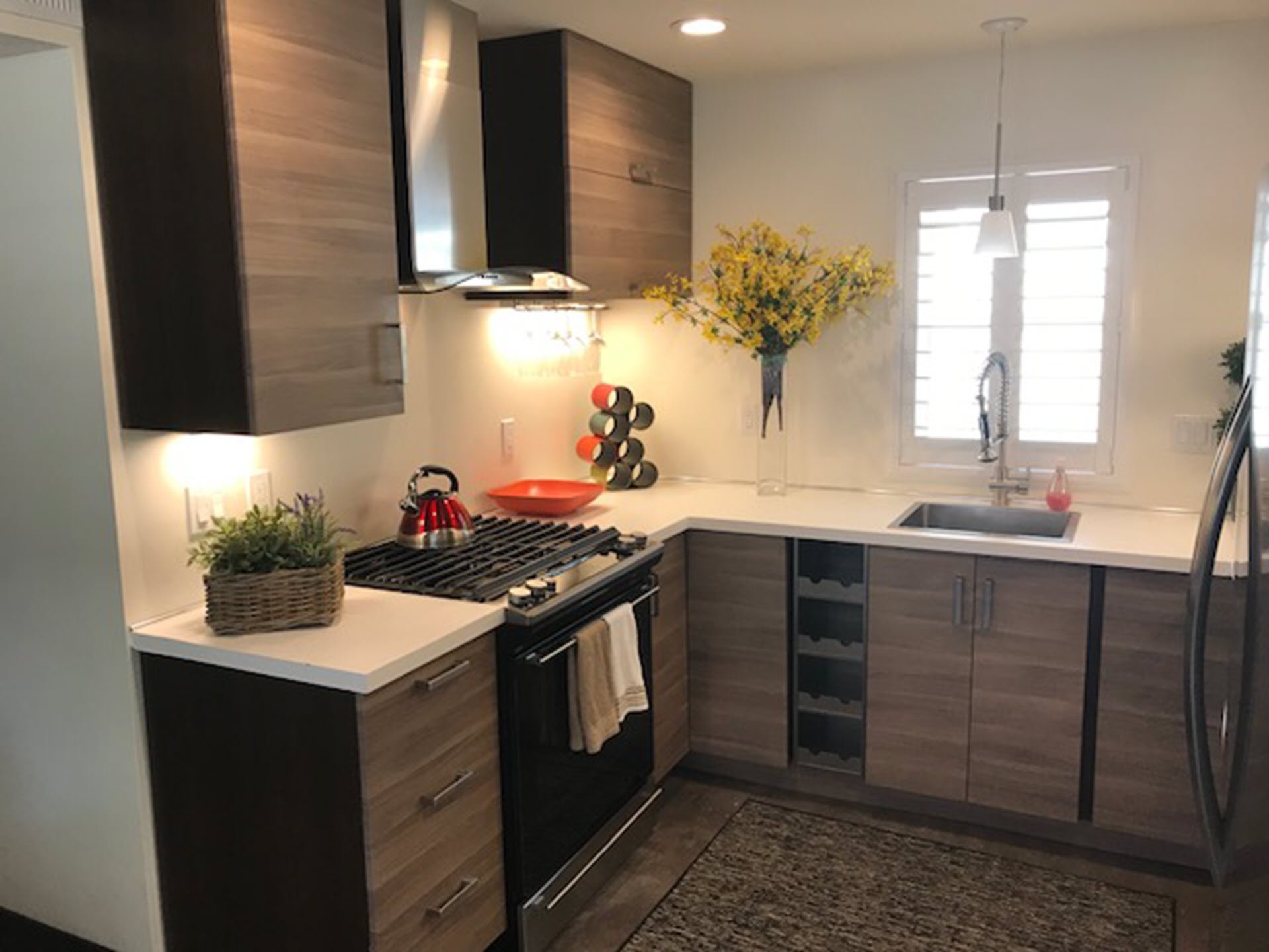 Modern U-shaped kitchen with wooden cabinets, white countertops, a stainless steel stove and oven, a red kettle, a green potted plant, a vase with yellow flowers on the countertop, a window with white shutters, and a pendant light hanging over the sink.