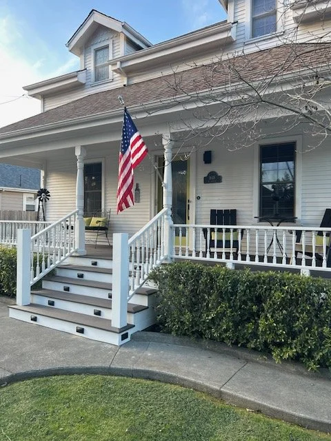 Front porch of a white house with an American flag, two black chairs, a small table, and decorative elements, with a landscaped front yard and a curb.