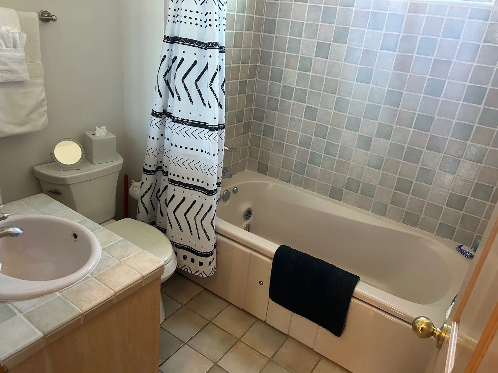 A small bathroom with a bathtub against a wall tiled with light-colored square tiles, a white shower curtain with black geometric patterns, a white toilet with a small mirror and tissue box on top, and a sink with a beige tiled countertop. A black towel hangs over the side of the tub, and a red plunger is behind the toilet.