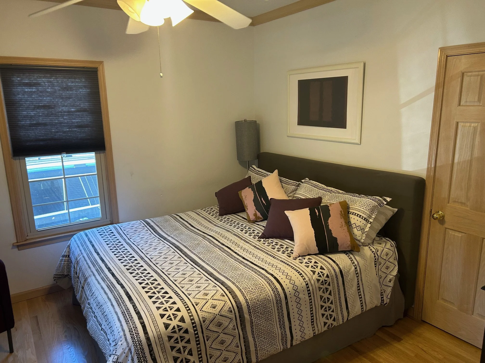 A bedroom with a bed covered in a patterned black and white bedspread, four decorative pillows on top, a window with a dark blind, an ceiling fan, a wall-mounted lamp, and a framed empty picture above the bed. There is a wooden door to the right and wooden window trim.