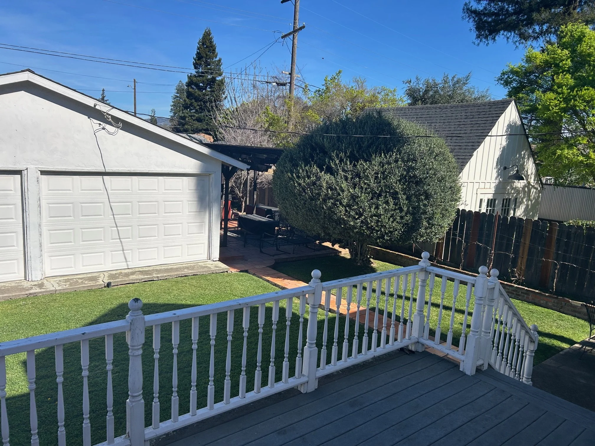 View of a backyard with a white picket fence, a grassy area, a large bush, a garage, and a small white building with a gabled roof under a clear blue sky.